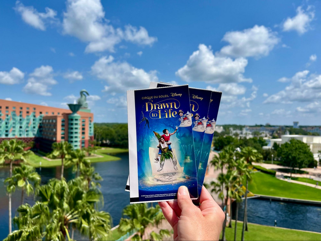 A hand holds Cirque du Soleil performance tickets. In the distance is the architecture of the Dolphin hotel, part of the legendary Walt Disney World Swan and Dolphin Resort. 
