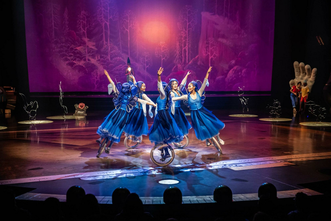 Five Cirque du Soleil performers dressed in blue fairy costumes ride unicycles in a clockwise circle, touching hands in the middle with their other hands outstretched upward. 