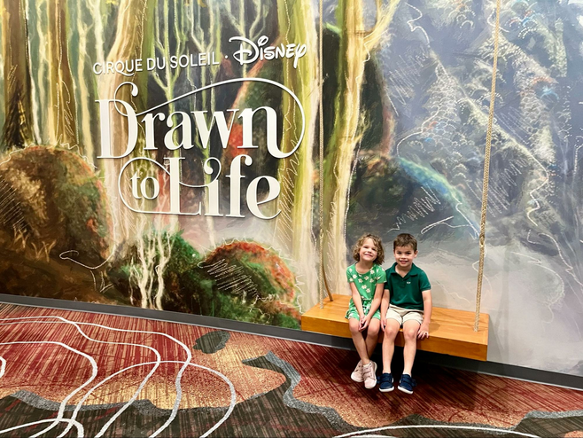 Two children smile in front of a forest-themed Cirque du Soleil backdrop.