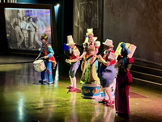 Five musicians in eclectic, colorful costumes play music onstage during a Cirque du Soleil performance. Behind them is a projected photo of The Animators, who are characters in the show. 