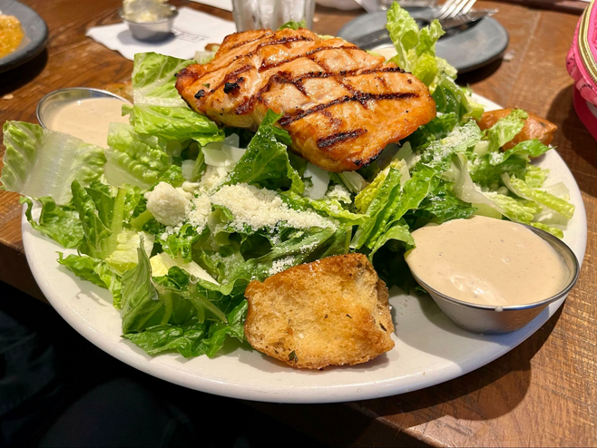 A piece of grilled salmon sits atop a Caesar salad, with two silver containers of salad dressing on the side.
