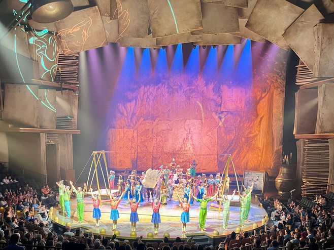 On a stage lit by blue, orange and purple stage lights, costumed Cirque du Soleil performers stand with their hands raised in the air during final bows.