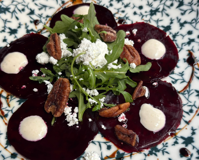Five beet slices arranged on a plate and topped with candied peans, goat cheese, arugala, and a dressing.