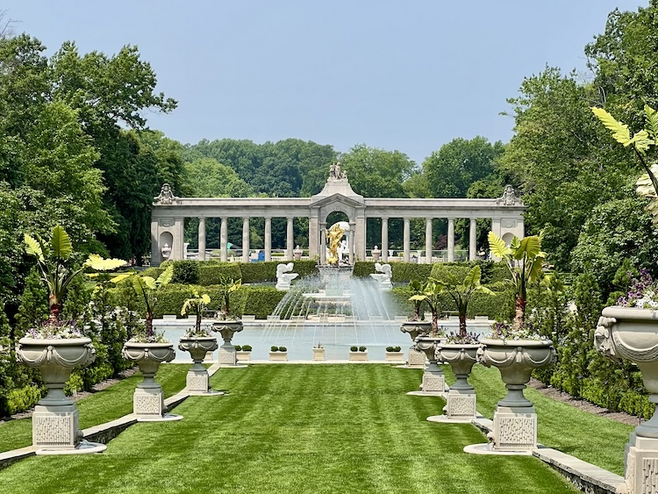 Formal French gardens at Nemours Estate, featuring fountains, ornamental urns, and neatly manicured greenery framed by surrounding trees.