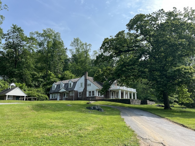 Classic painted white wood and brick home set beneath towering trees with a rolling green lawn in the foreground.