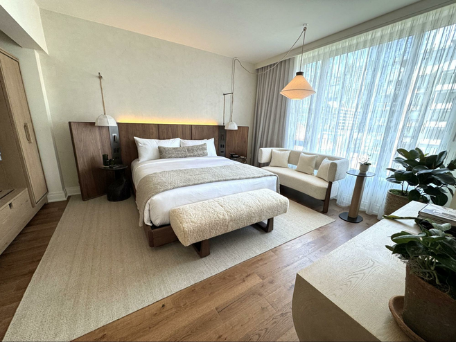 hotel bedroom with neutral colored linens and rugs with natural wood accents, bed, loveseat and dresser with hanging lamps