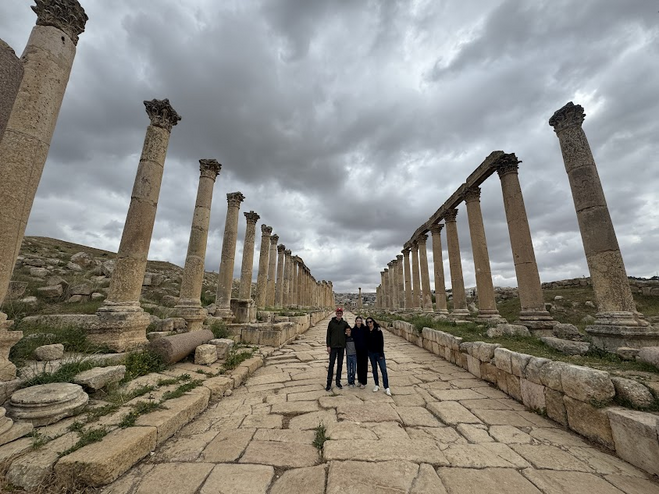 Father in red hat, mother in black sweater, a boy and a girl among the Roman ruins in Jerash. There are tall columns.