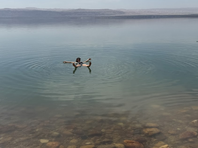 Woman wearing sunglasses floating in sunglasses floating in the Dead Sea in Jordan. Brown rocks are in the foreground and the water is blue. Mountains are in the background.