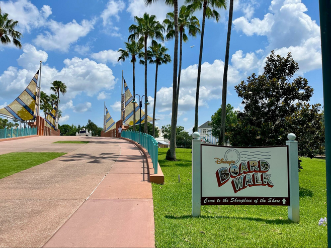 A paved coral-hued walkway that passes over a canal to Disney's BoardWalk, lined with palm trees and a sign for Disney's BoardWalk pictured on the right.