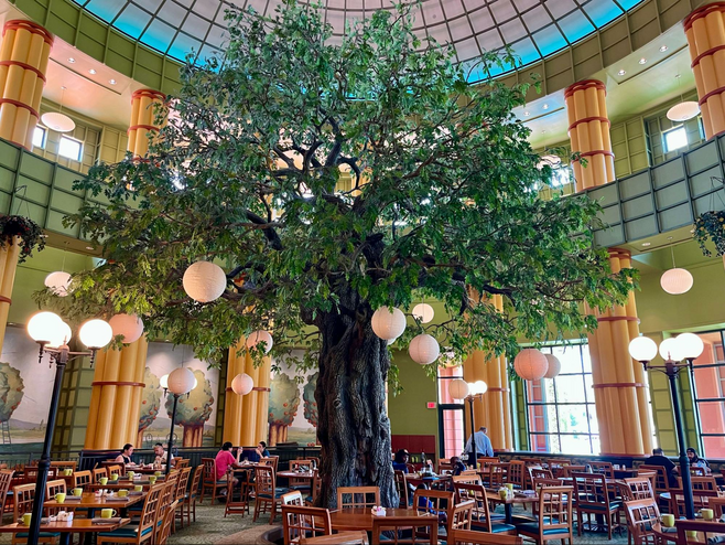 A giant faux oak tree stands in the center of a high-ceiling restaurant. The space has wooden tables and chairs (with some patrons eating), large windows and is filled with natural light. 