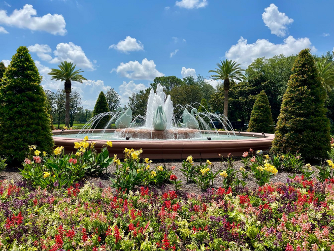 A circular outdoor fountain features three swan sculptures and water spouts. It is surrounded with beautifully-landscaped red and yellow flowers and decorative trees.