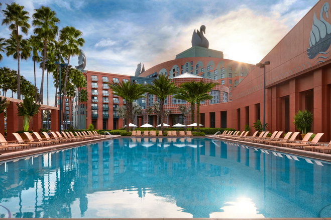 A swimming pool is framed by a coral-and-turquoise hotel building, palm trees and deck chairs. The building has a giant swan sculpture on its roof. 
