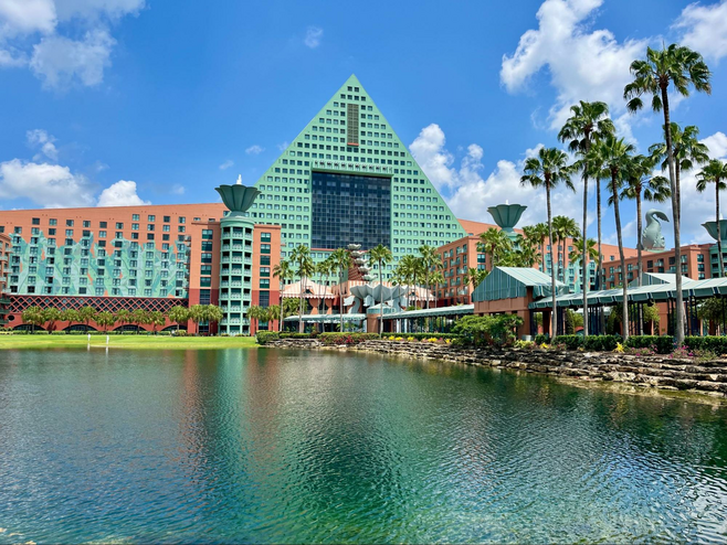A coral-and-turquoise hotel with a triangular center is pictured across a waterway, with a covered walkway visible to cross.