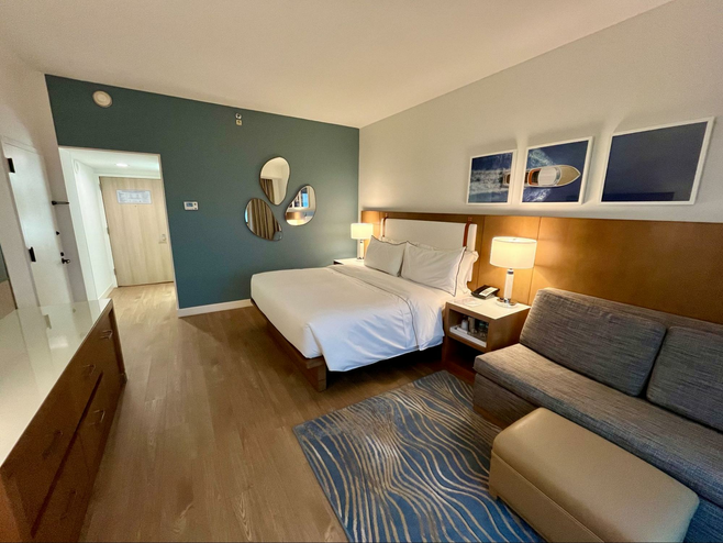 A view of a newly-renovated bedroom. Pictured are a modern wood king-sized bed, a modern wooden dresser with a white glass top, a taupe sofa and three minimalist decorative mirrors.