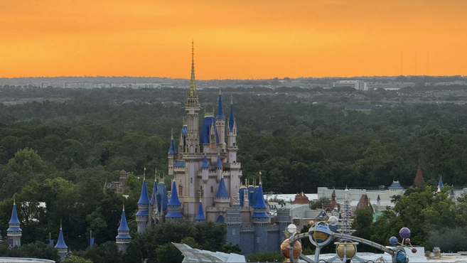 Orange yellow sky for sunset with tall Cinderella castle in the forefront with greenery in the background.