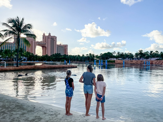 Three pre-teen and teen girls stand at water's edge on the beach with palm trees and the Atlantis towers in the background.
