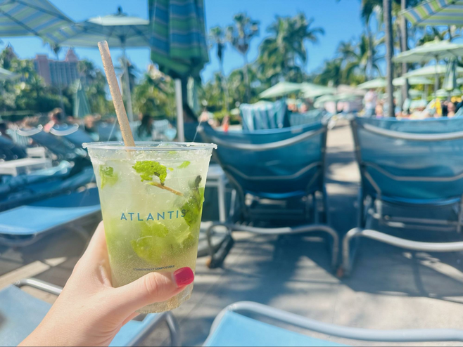A female hand with pink thumbnail holds up a cocktail with green leaves and a straw sticking out with a pool deck in the background.