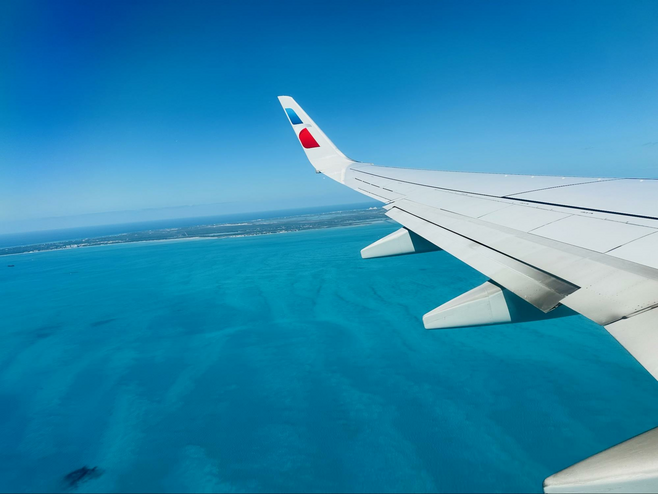 View from an airplane with a portion of the wing visible. Below is blue water with an island in the distance.