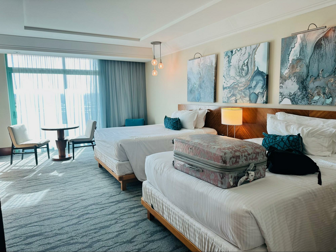 View of large two-queen hotel room with small round table and two chairs behind the beds and a suitcase and tote on the closer bed. Sheer curtains cover a slider down at the back of the room with sunlight filtered inside.