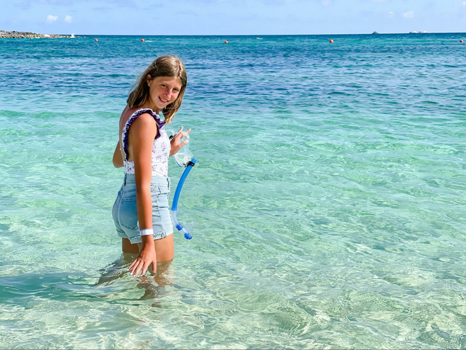 A young girl in bathing suit and denim shorts stands in the turquoise water at Atlantis, with her head turned toward the camera, with one hand dipped into the water and the other hand holding a snorkel mask.