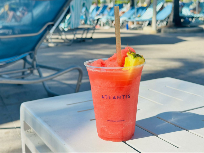 A pink smoothie-like beverage with a piece of pineapple and a straw sticking out, sits on a chairside table on a pool deck. The clear plastic cup says 'Atlantis'.