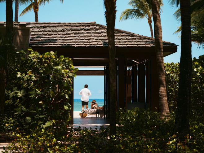 View of a cabana at Atlantis Paradise Island in the Bahamas. Through the doorway you can see a man standing in a white shirt and dark swim trunks and a woman lying on a beach chair, with the ocean in the background.