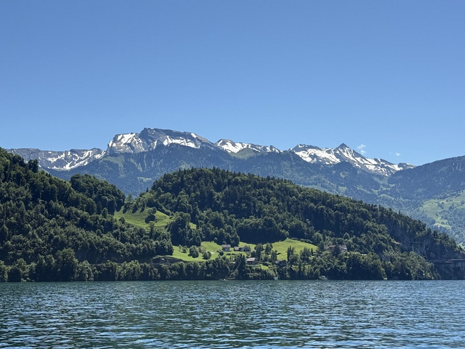 Snow capped mountains overlook rolling hills with green trees along a large body of water