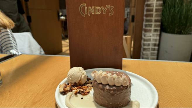 Chocolate cake on top of a white plate infront of a brown menu with the word "Cindy" written across. 