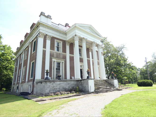 A two story Greek revival mansion with white columns in front with a curved driveway in front.