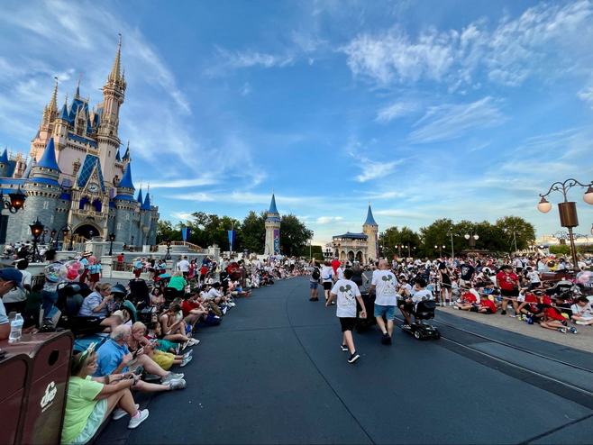 Crowds of people are seated on either side of a parade route at Disney's Magic Kingdom, with Cinderella's Castle pictured on the left side behind them.