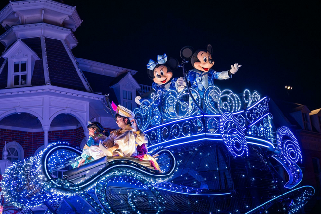 Costumed characters Jasmine and Aladdin sit on a lit-up parade float shaped like a Magic Carpet, while Minnie and Mickey Mouse wave from a perch above. All are on a glittering parade float at night.