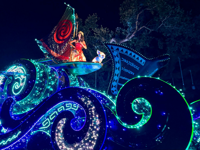 A performer dressed as Moana is perched on a float meant to look like a boat sailing on glittering waves. The float is covered with sparkling lights, and the photo is taken at night, so everything shimmers.