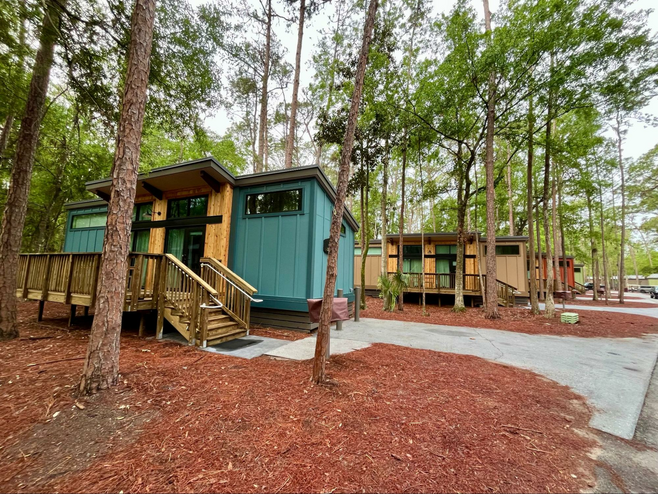 Modern cabins are pictured in a forest, with paved walkways and steps leading up to each porch.