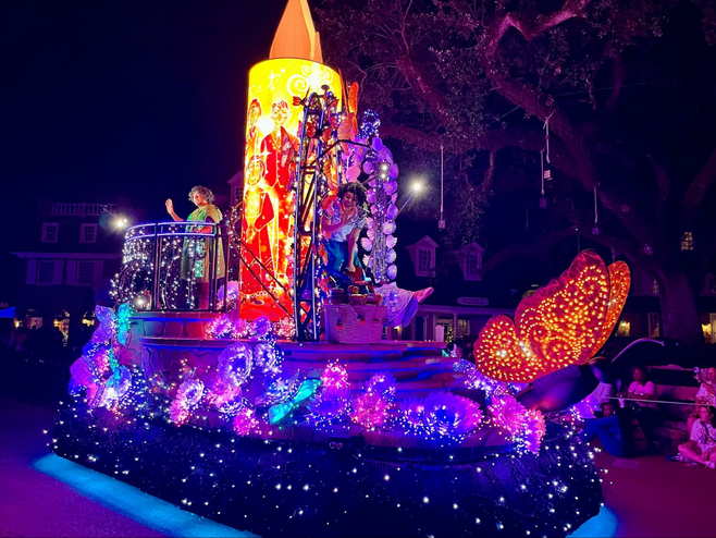 Actors dressed as Bruno and Mirabel from Disney's film Encanto smile and wave from a sparkling parade float. The float is covered in tiny colorful lights, and the photo is taken at night, so the float shimmers.