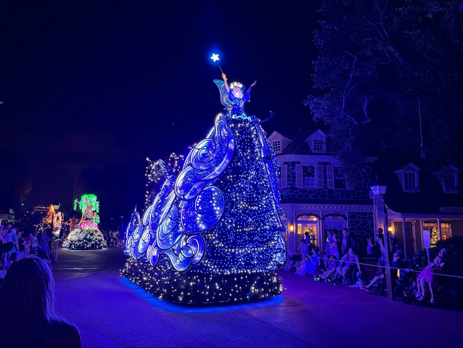 A performer is perched atop a high float covered in glittering lights during an evening parade. The performer is dressed as the Blue Fairy from Pinocchio and is holding a wand shaped like a star high in the air.