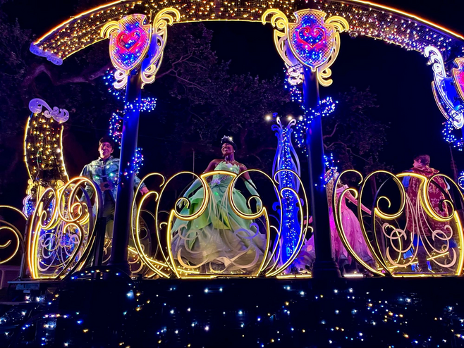 A prince and princess dance on a float covered in glittering, colorful lights during a nighttime parade. The princess is wearing a beautiful green ballgown.