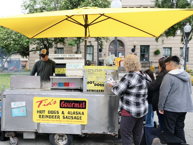 Anchorage food cart with yellow umbrella and customers lined up for reindeer sausage 