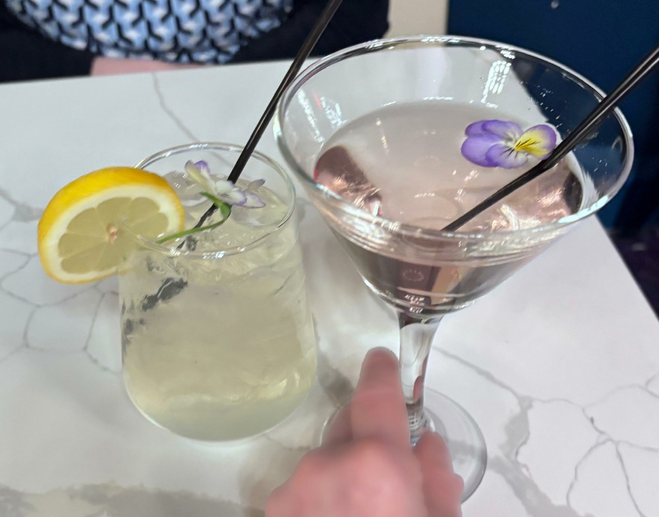 A small glass with a clear cocktail and an orange slice wedged on the glass and a martini glass with pinkish liquid and an elderflower. 
