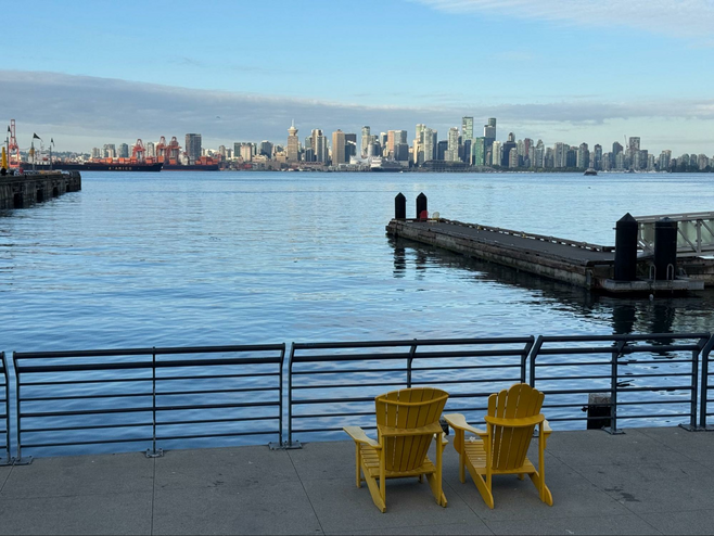 Two yellow adirondack chairs overlook the waterfront from a pier with the city of Vancouver in the distance.