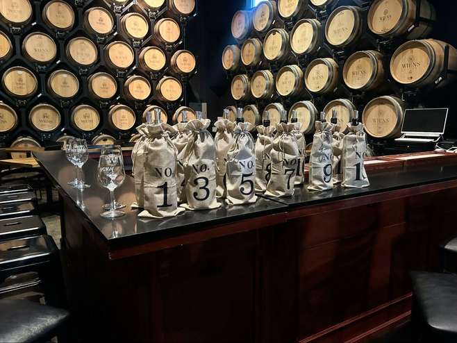 Wine bottles covered in opaque sacks line the serving counter inside the barrel room. In the background are barrels stacked on top of one another in an orderly fashion at the Wiens Cellars in Temecula, CA.