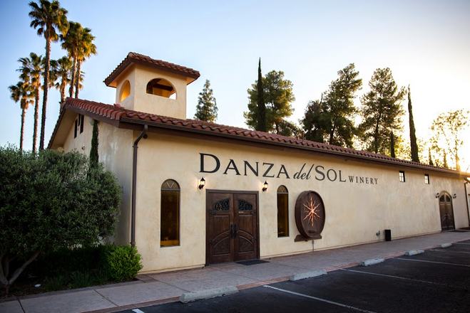 exterior of building that reads "Danza del Sol Winery". Building is biege with brick rooftops and towering trees peering over the building