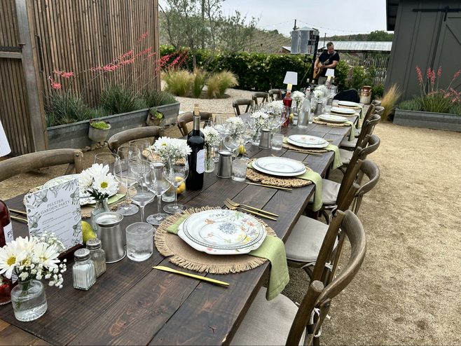 An elongated outdoor dining table at the Peltzer Winery is set for a wine tasting, with wine glasses and dinner sets along with decorative flowers. In the background, a guitarist warms up.