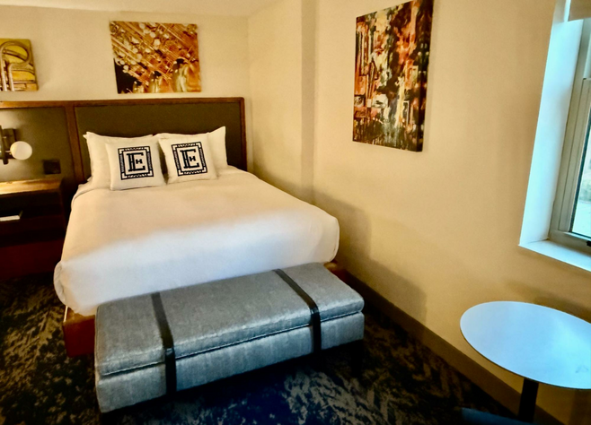 Queen-size bed with two pillows bearing a big letter "E"  for Elkhart has a grey bench at the foot, the width of the bed. Walls are a calm yellow-beige.