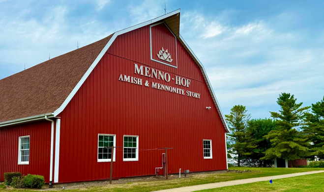 Blue sky with wisps of white clouds frames the red barn. Windows suggest only one floor inside but the roof towers as if several stories. Big letters easy to see from the road say MENNO-HOF: Amish & Mennonite Story.