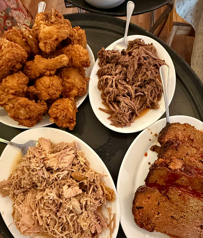 Crispy fried chicken legs, thighs and breasts are piled on top of each other on a platter next to shredded pork. The pork plate touches a platter of meat loaf slices big enough to be shared. In the lower corner of the serving tray is turkey.