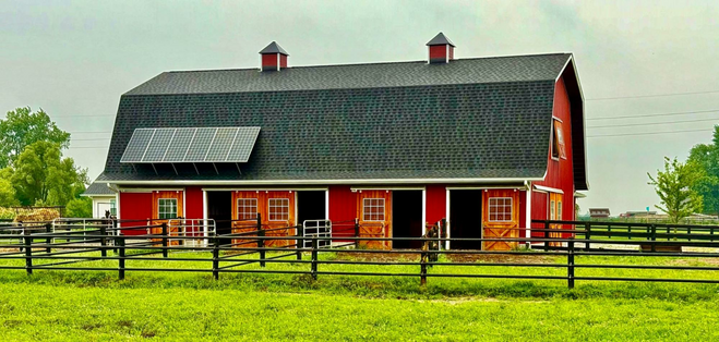 Red barn with four doors to horse stalls holds six solar panels on one side of the steep, sloping black roof.