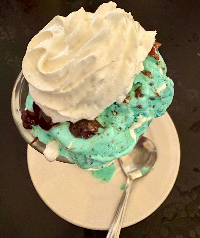 Green chocolate chip mint ice cream oozes over the edges of the tall glass sundae dish which is barely visible. A swirly dollop of whipped cream melts on top of the ice cream.