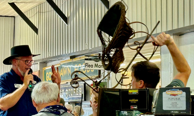 Auctioneer wearing a broad-brimmed black Amish hat holds up his left-hand  pinky, ring and middle fingers while his right hand holds a microphone. Helper hoists a child-sized sleigh over his head so shoppers can call out their bids. 
