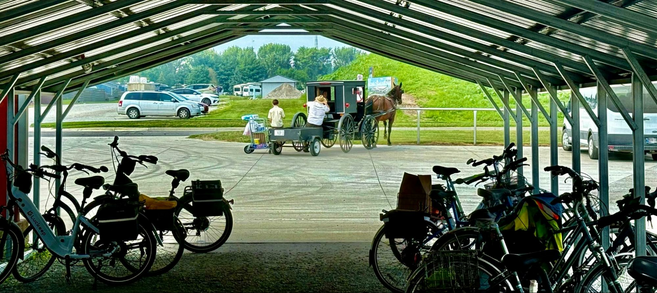 Outside a grocery store, bicycles with saddlebags are parked under an open-sided, metal-roofed shed. In the foreground two men load groceries into their black buggy while the horse is tied to a rail. A small cart is fastened to the back of the buggy.