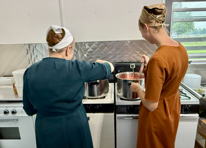 Grandmother on the left, shorter than adult granddaughter on her right: both stirring tall-sided pots to make jelly. Three four-burner stoves are side-by-side in the small kitchen. The women wear simple straight dresses with elbow-length sleeves. Triangle scarves cover their hair, tied in the back.